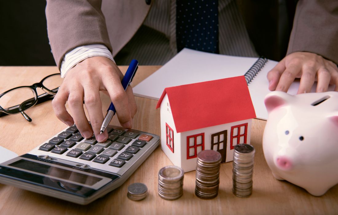 The image shows a person calculating home financing costs with a pen, surrounded by a toy house, stacks of coins, and a piggy bank, representing savings and budgeting for a home purchase.